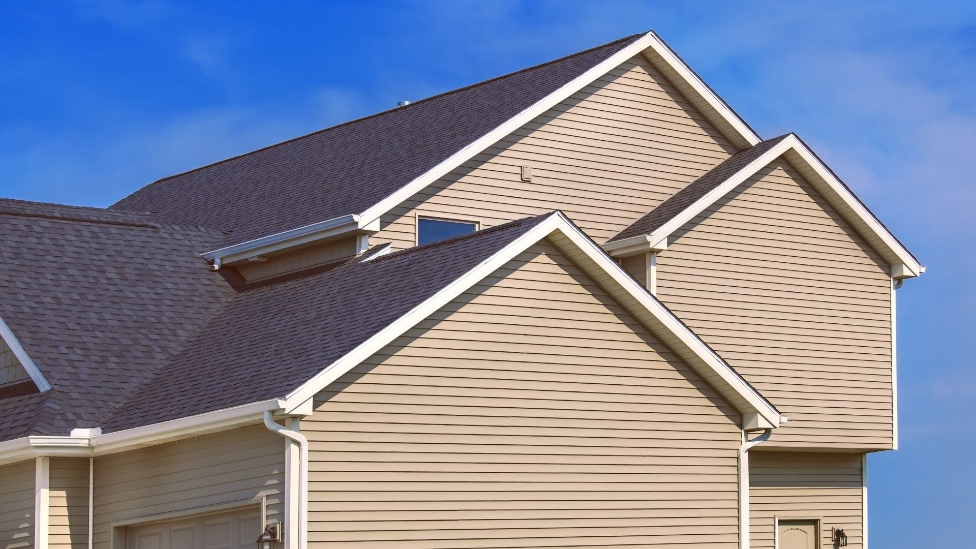 Beige house with multiple roof levels and dark shingles against blue sky